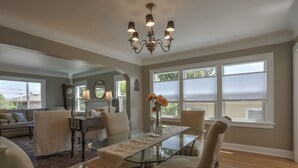 Uma área de refeições com mesa de vidro, cadeiras e um candelabro. Há janelas com persianas, um sofá e um abajur ao fundo.
