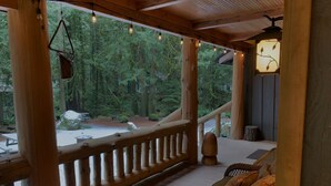A wooden deck with a wicker sofa and hanging lights, overlooking a forested area with a stone bench and a fireplace. -  Spring /Summer availability!  On 2 creeks, wood burning fireplc, log gazebo. (Rhododendron)