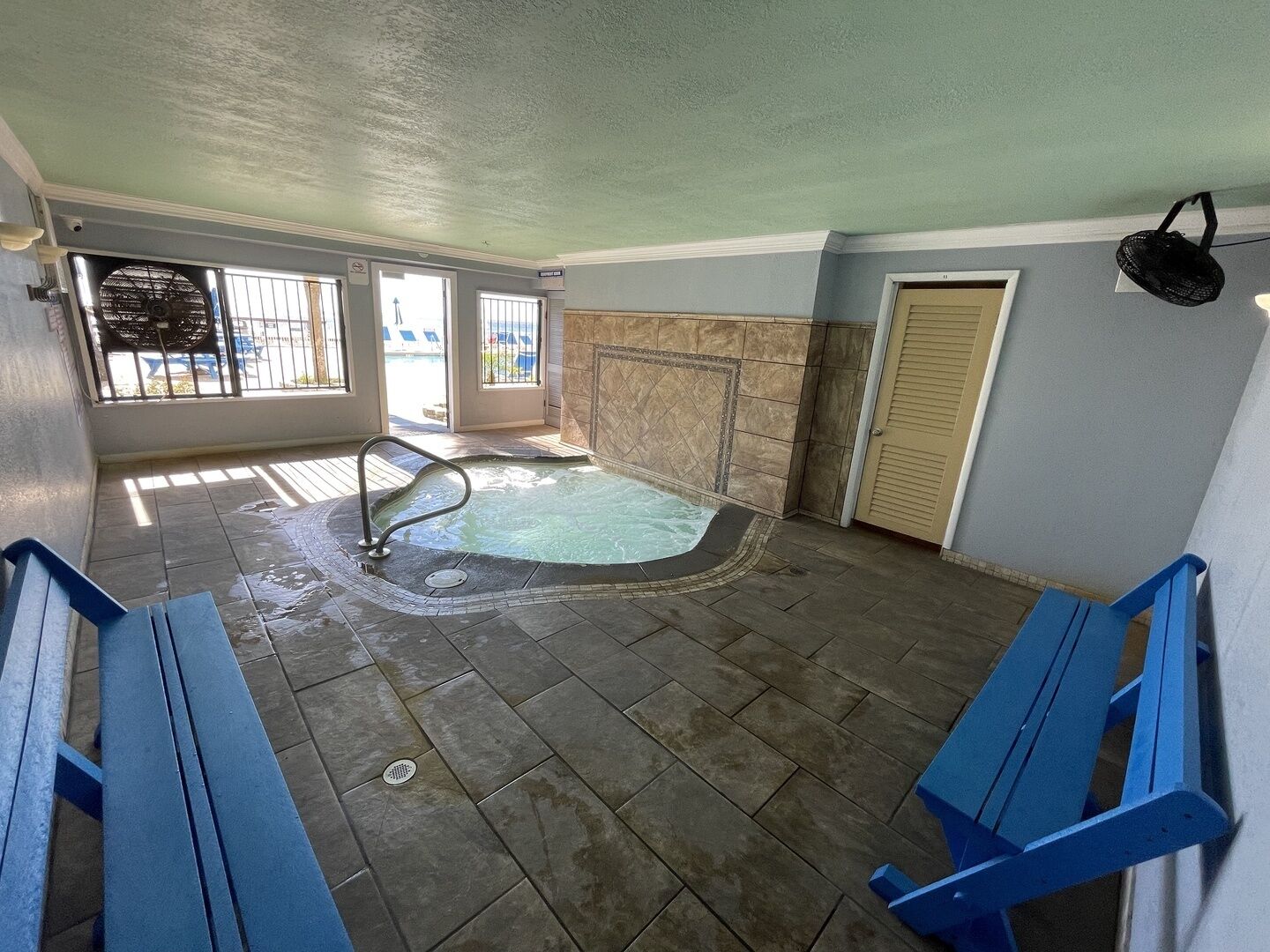 Et spaområde med jacuzzi, bænke og udsigt til en pool samt liggestole gennem vinduerne.
