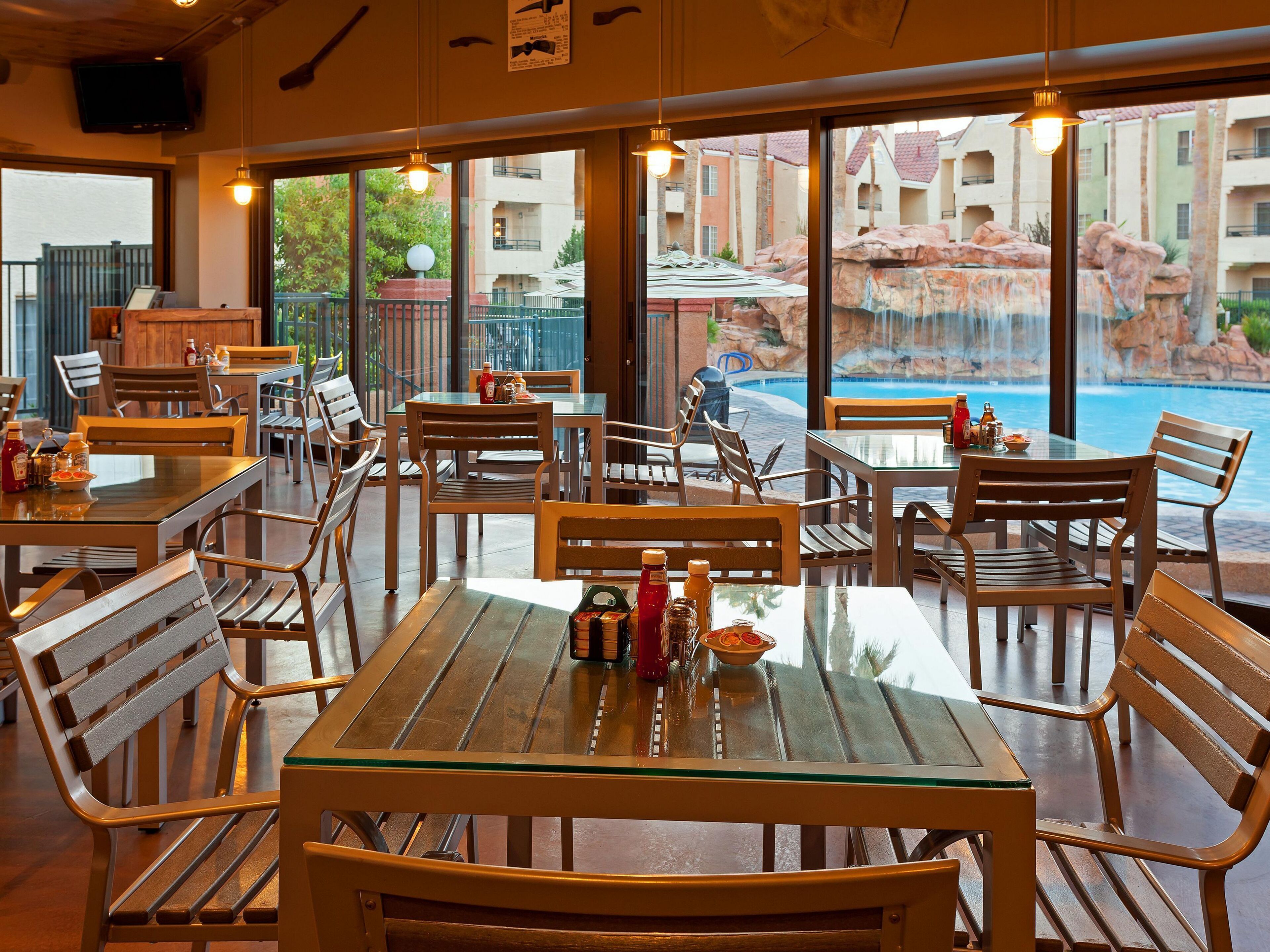 A restaurant with glass tables and wooden chairs, overlooking a pool with a waterfall and buildings in the background.