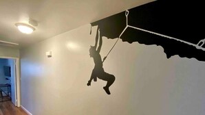 A room with a wooden floor, a white wall, and a silhouette of a person climbing a wall.