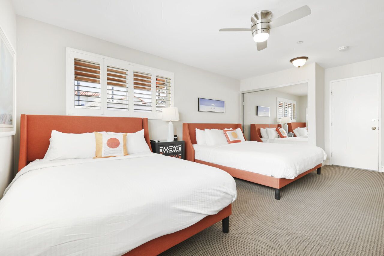 A hotel room with two beds, a ceiling fan, a window with blinds, a bedside table with a lamp, and a framed picture on the wall.