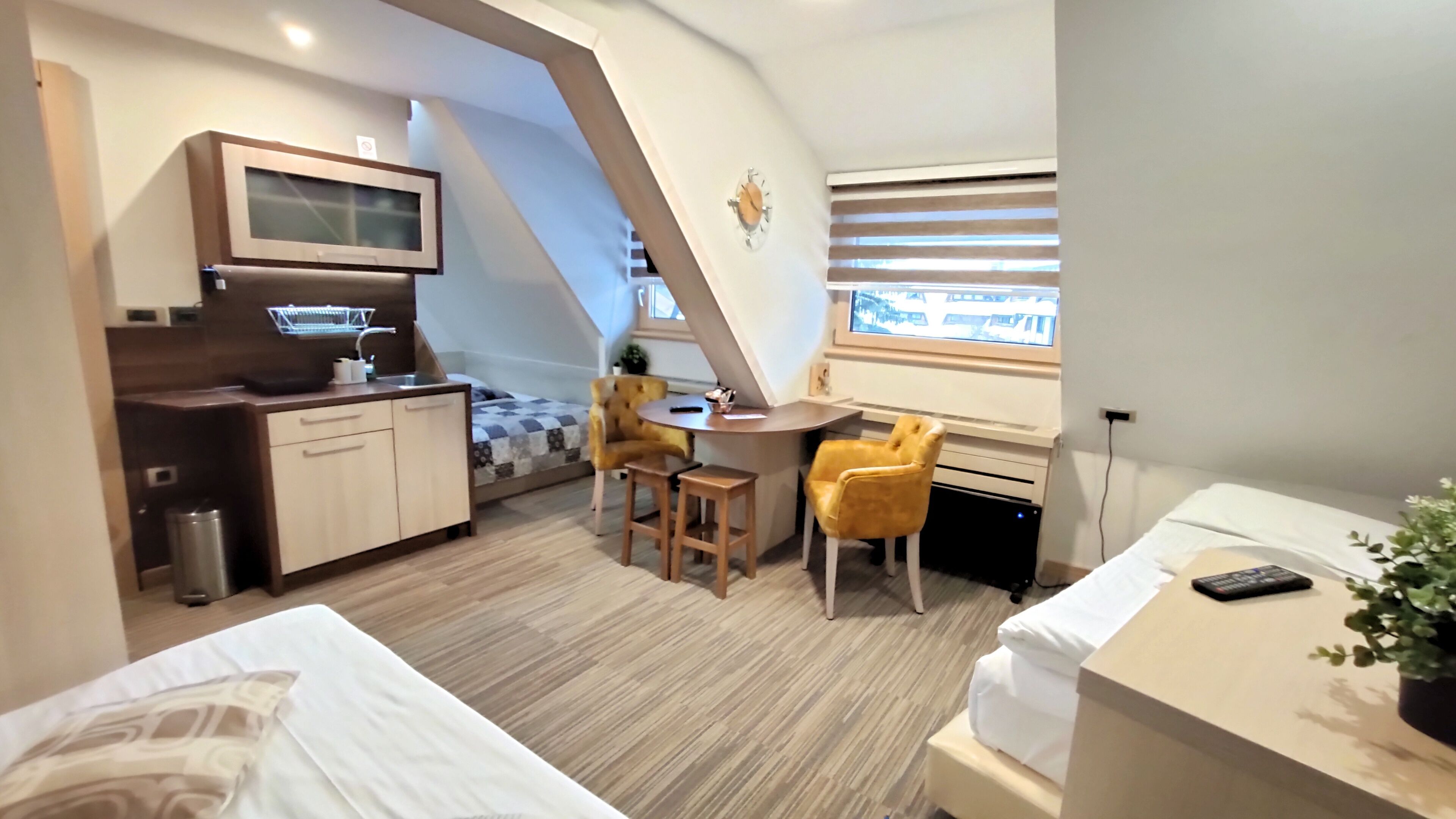 A compact hotel room with a kitchenette, a bed, a small dining area, and a window with blinds.