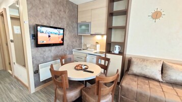 Un espacio habitable compacto con zona de comedor, kitchenette y un televisor mostrando un partido de tenis.