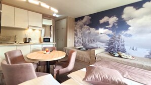 Moderni hotellihuone, jossa on keittotila, hedelmillä koristeltu ruokapöytä, koristeellisilla tyynyillä varustettu sohva ja seinämaalaus lumisesta maisemasta.