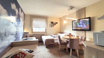 Una habitación de hotel acogedora con cama, sofá, mesa de comedor con sillas, televisor y un amplio ventanal con persianas.
