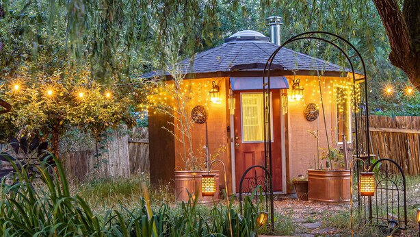 Un sentier en bois mène à un petit bâtiment accueillant, doté d’une porte rouge, et entouré d’une végétation luxuriante et de guirlandes lumineuses.