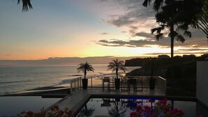 Área de piscina com vista para o mar ao pôr do sol, mobiliário externo e palmeiras.