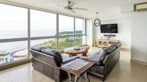 Uma sala de estar ampla com uma grande janela com vista para a praia, um ventilador de teto, uma televisão de tela plana e uma mesa de centro de madeira.