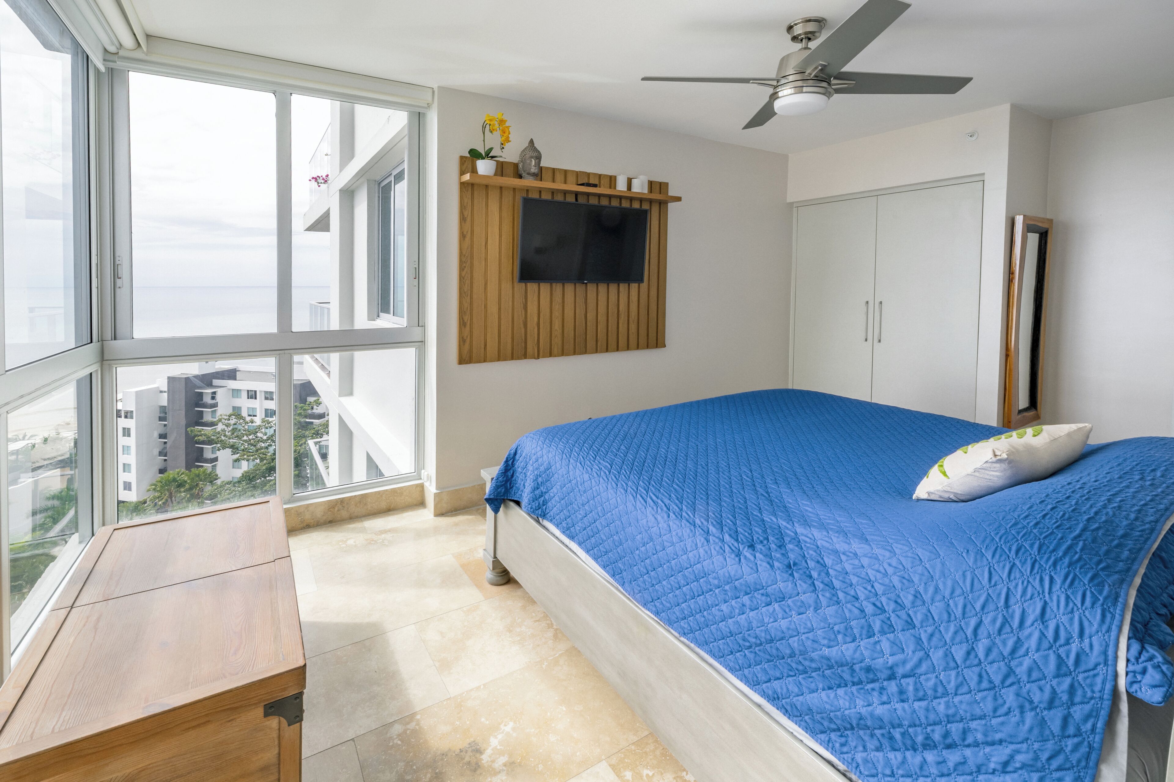 Une chambre avec une grande fenêtre, un lit recouvert d’une couette bleue, un ventilateur de plafond et un téléviseur fixé au mur.