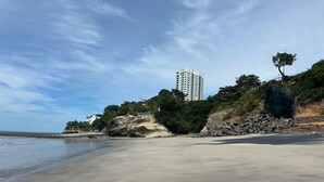 Uma praia com areal, um afloramento rochoso e um edifício alto ao fundo.