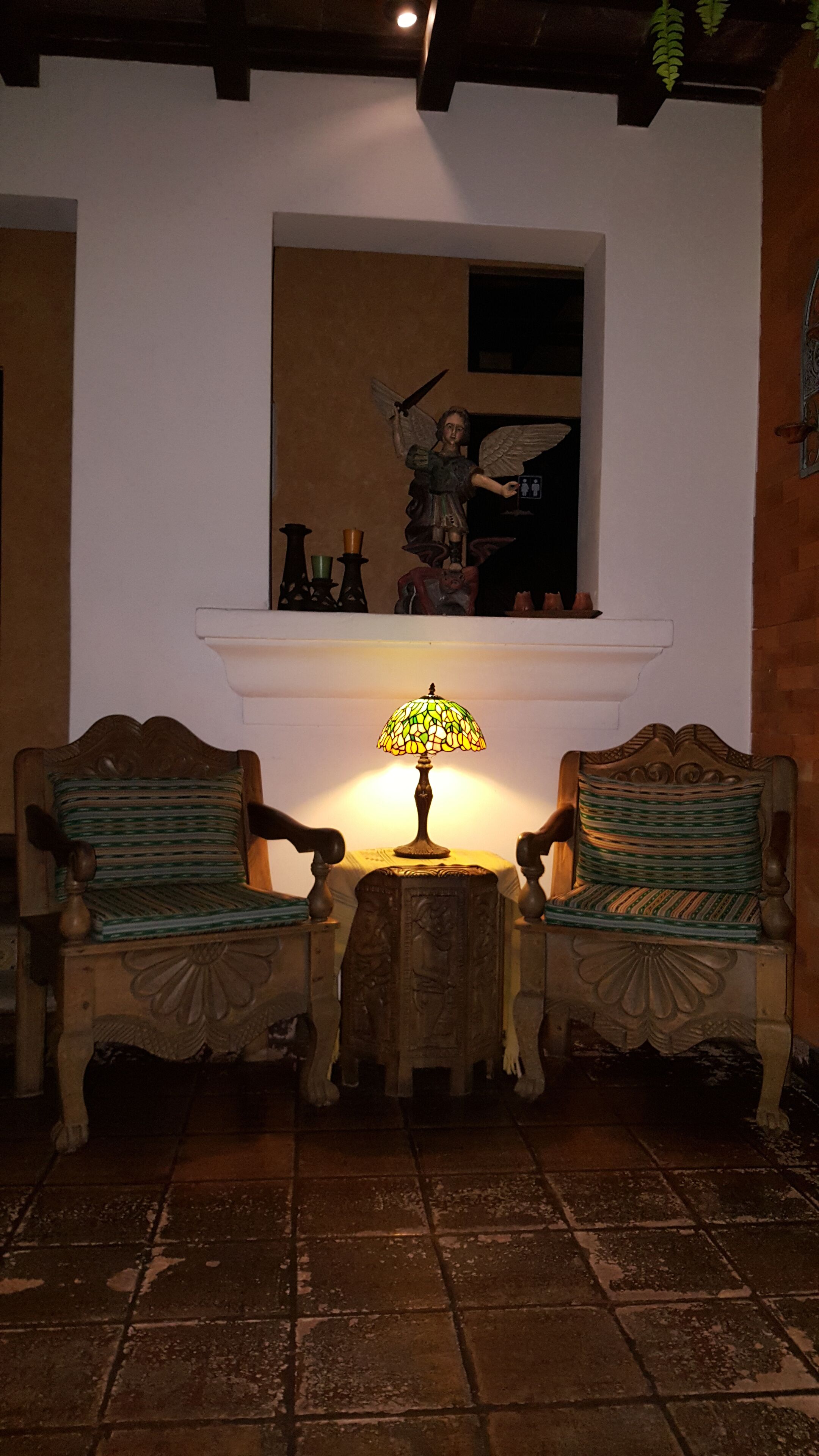 Una habitación con dos sillas de madera, una mesita con una lámpara de vitral y un estante de pared con adornos.