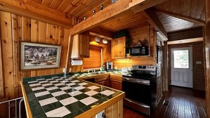 Interior de una cabaña de madera con cocina, que cuenta con una encimera de baldosas a cuadros, armarios de madera, una estufa y un microondas.