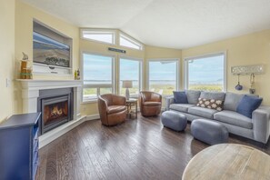 A living room with a fireplace, leather chairs, a gray sofa, and large windows offering a view.