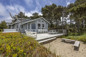 A coastal house with a wooden deck, white railings, and a sandy beach in the foreground.