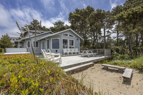 Une maison situĂ©e sur la cĂŽte, dotĂ©e dâune terrasse en bois, de balustrades blanches et dâune plage de sable au premier plan.