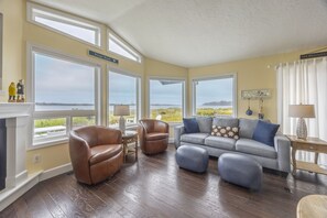 A living room with a fireplace, leather chairs, a grey sofa, and large windows with a view of the beach. - Waterfront | Hot Tub | Kayaks | Crab Traps | 3~Kings (Lincoln City)