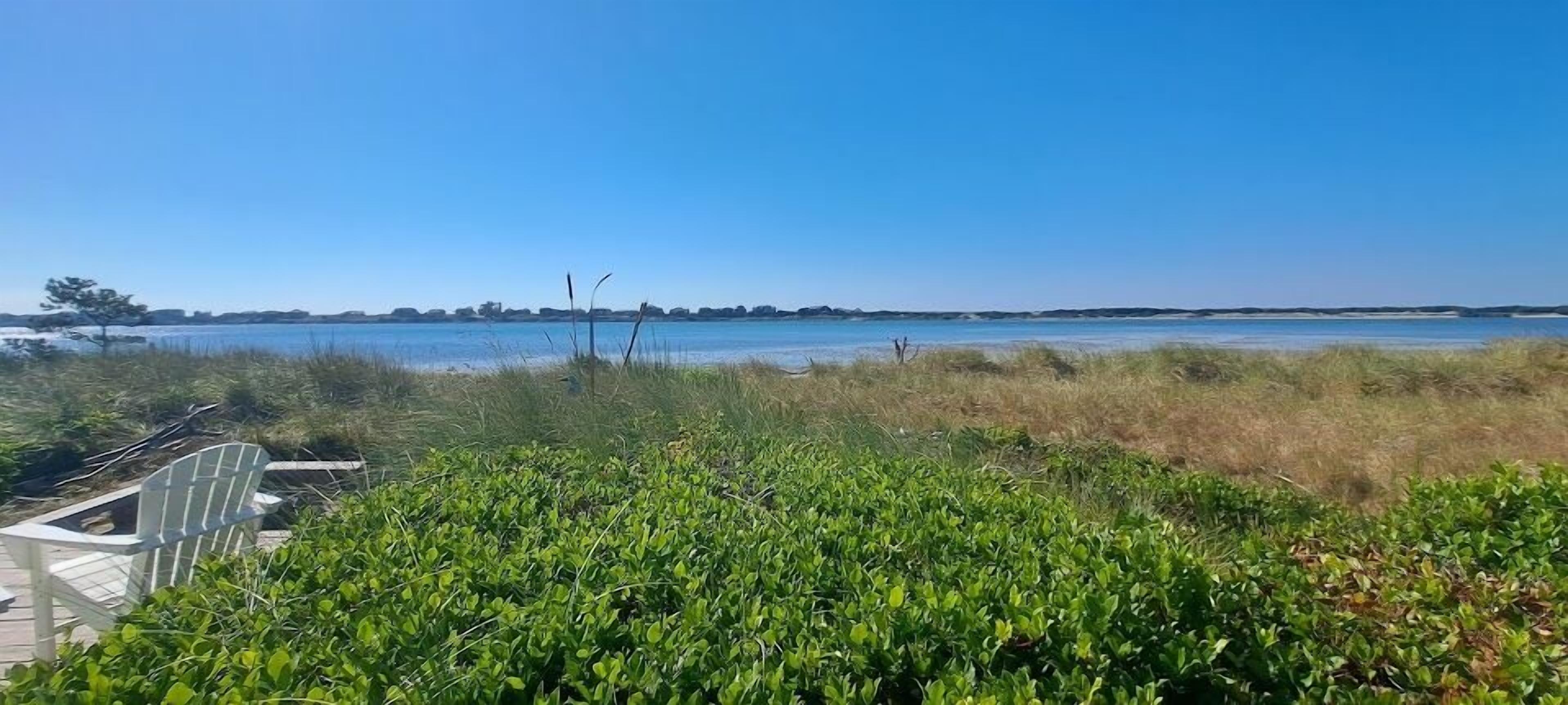 A beach view with a wooden deck and a chair, surrounded by green vegetation and a clear blue sky.