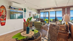 Habitación de hotel moderna con un amplio ventanal que ofrece vistas al mar, un área de comedor con mobiliario de mimbre y un televisor en la pared.