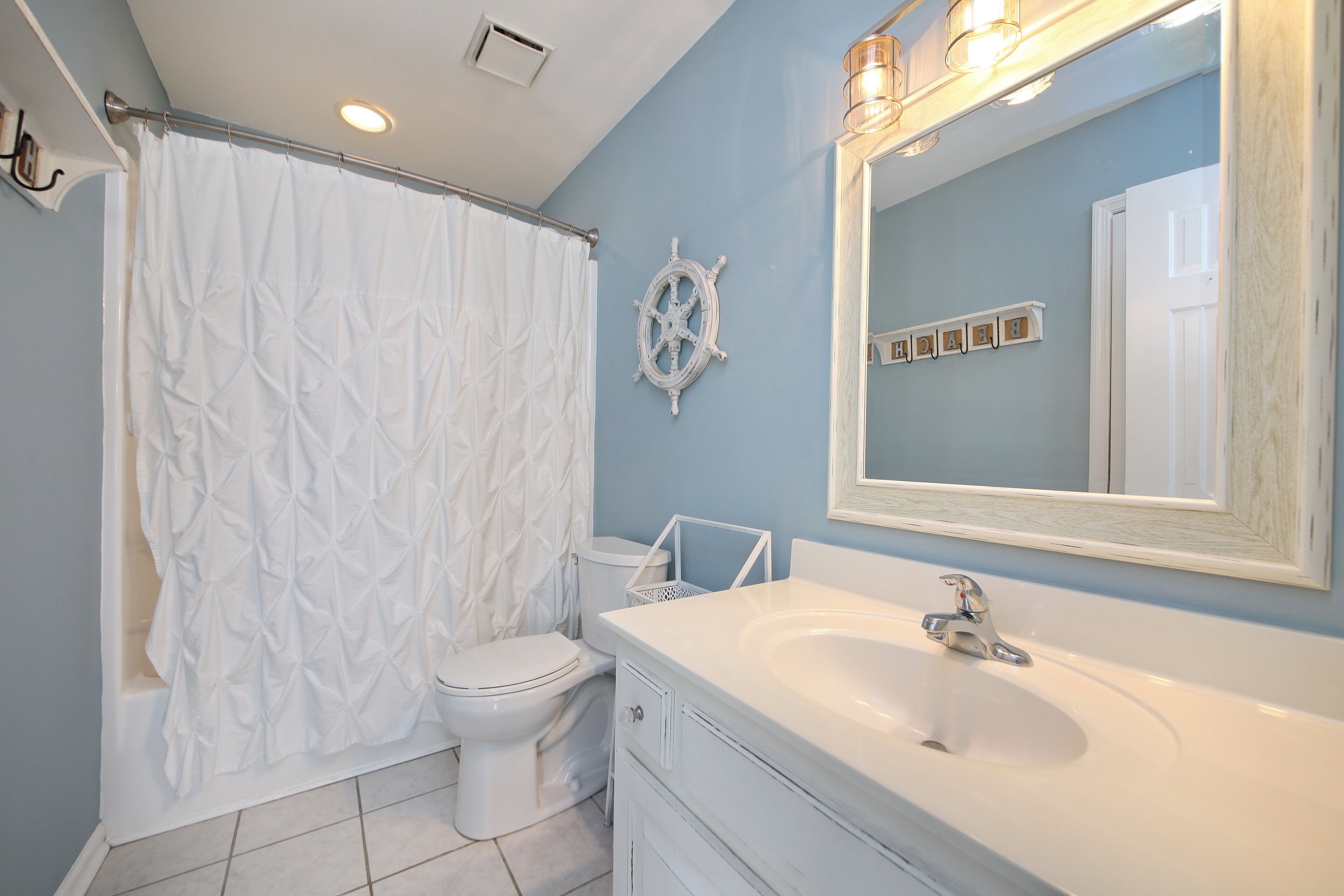 A bathroom with a white bathtub, a white toilet, a white sink with a mirror, and a ship's wheel decoration on the wall.