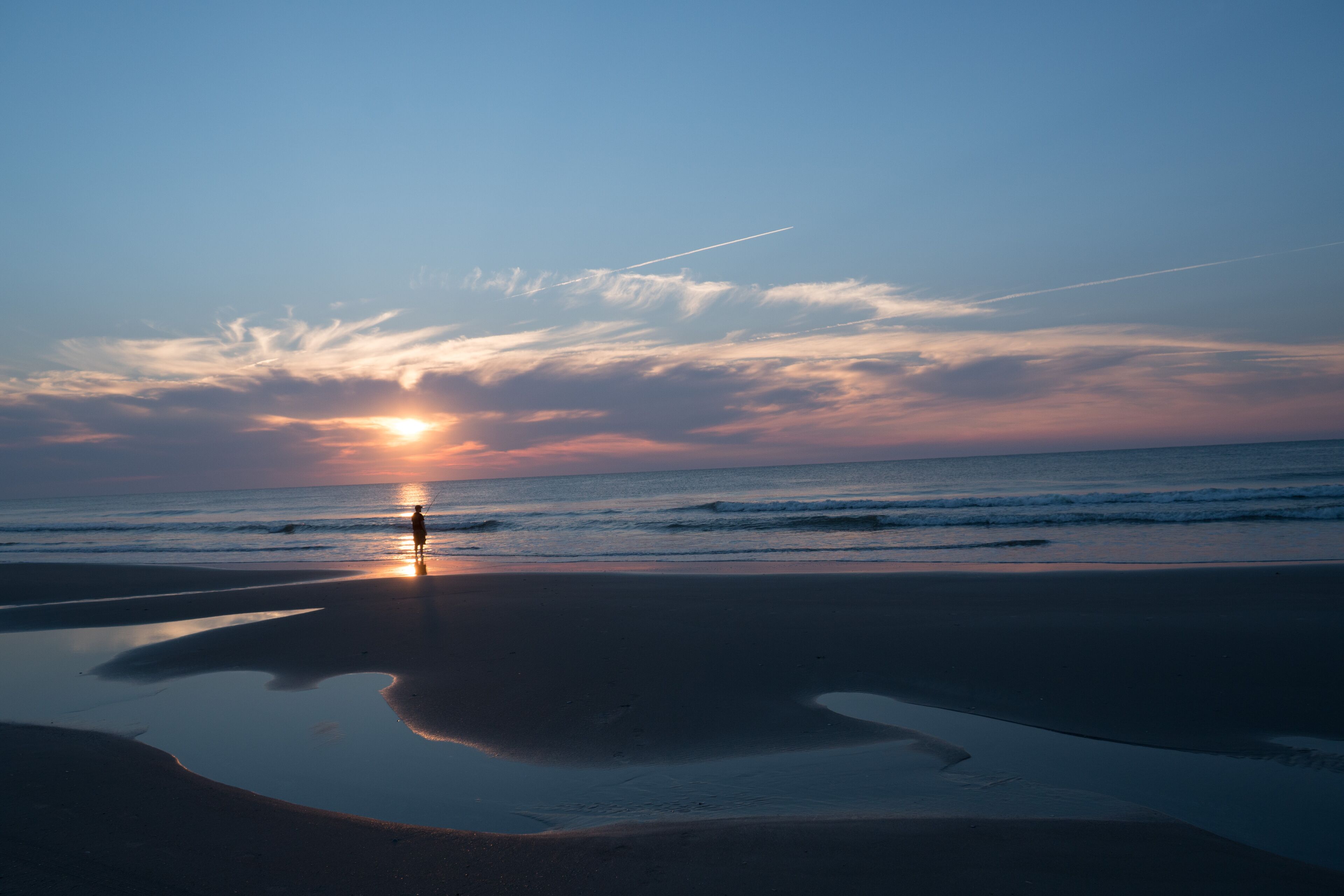 Cena de praia com uma pessoa de pé perto da beira da água, refletindo o pôr do sol na areia molhada.