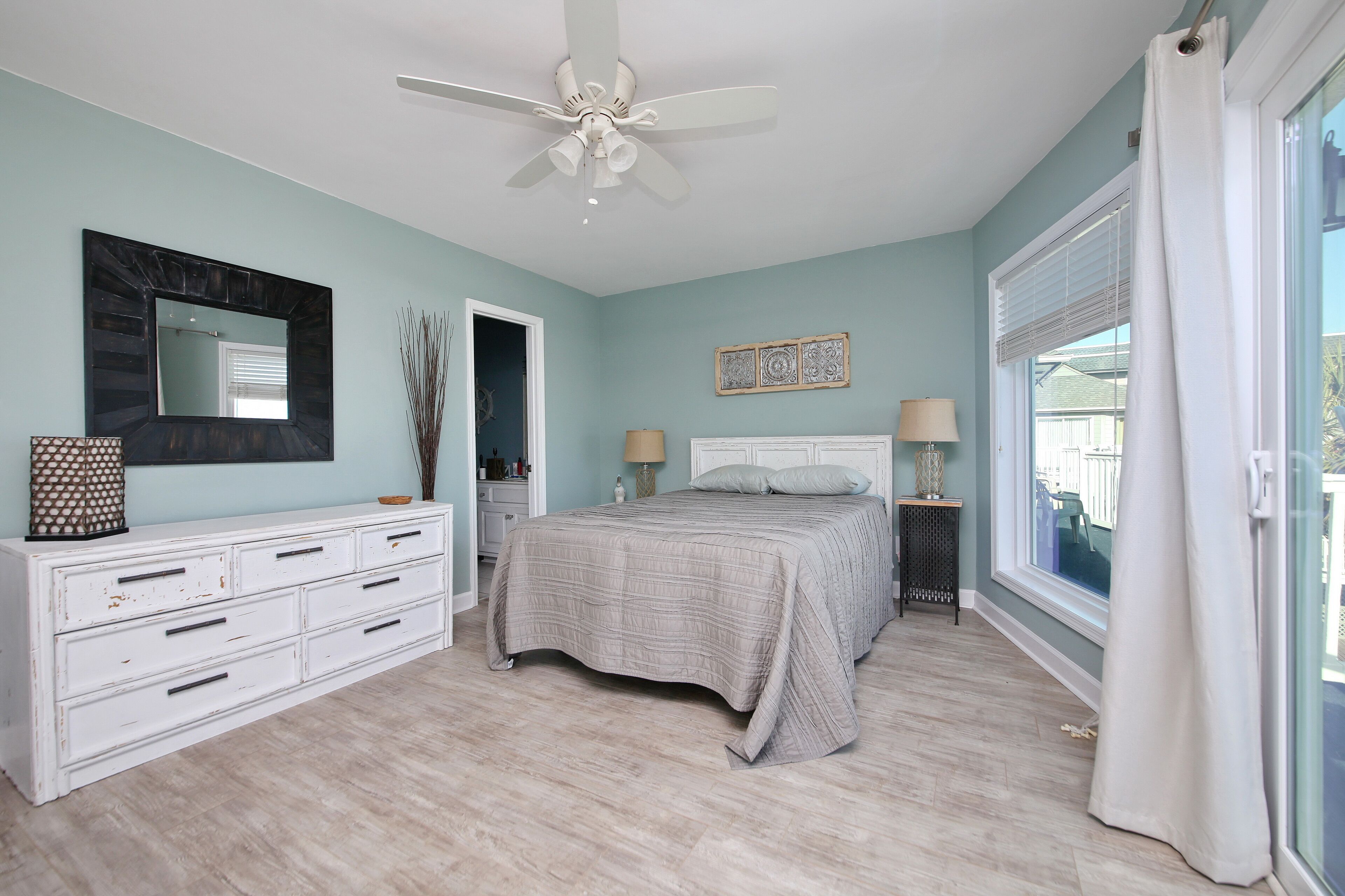 Un dormitorio con cama, cómoda, ventilador de techo y ventana grande con cortinas.