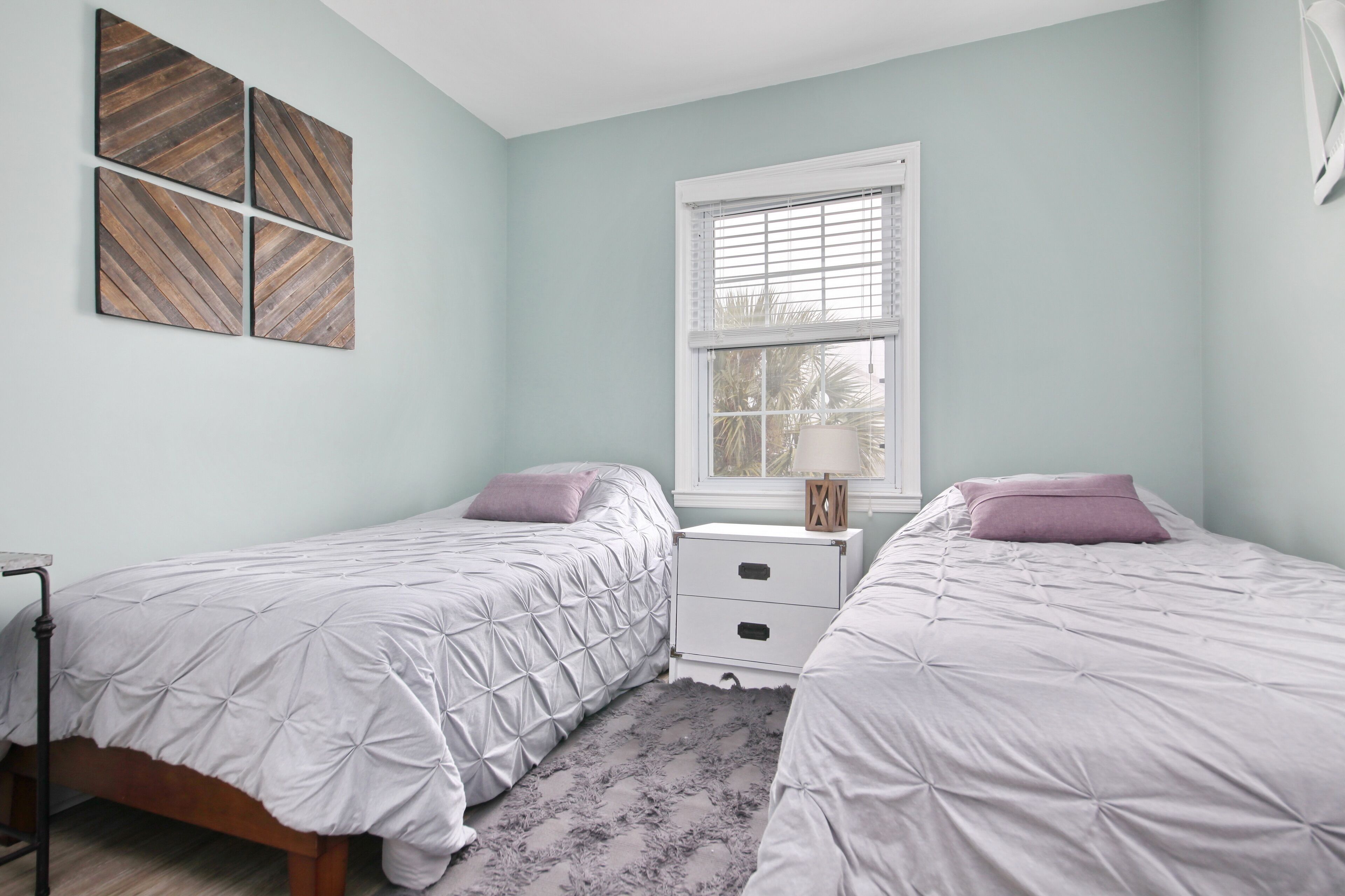 A bedroom with two beds, a window with blinds, a bedside table, and a wall-mounted artwork.