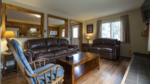 A living room with a brown leather sofa, a wooden coffee table, and a wooden rocking chair. There is a kitchen visible through an open doorway.