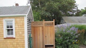 Un pequeño cobertizo de madera con techo de celosía y una puerta, junto a una casa con una ventana y una manguera.