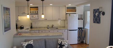 A compact kitchen with white cabinets, a microwave, a stove, and a refrigerator. There is a gray sofa with decorative pillows and a small table with a basket.