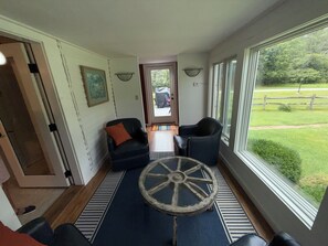 A room with a large window, a blue rug, two chairs, a small table with a wheel, and a door leading outside.