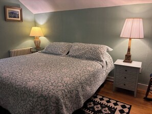 Quarto com cama, duas luminárias de cabeceira, mesa de cabeceira, cômoda, radiador e um quadro na parede.