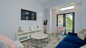 A modern living room with a round coffee table, a flat-screen TV mounted on the wall, a white console table with decorative items, a blue sofa, and a large window with blinds.