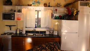 Een kleine keuken met een kookplaat, magnetron, gootsteen en koelkast. Er is een raam met gordijnen, een plankje met decoratieve objecten, en een bed met een beddengoed met een patroon.