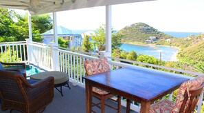 Un espace extérieur abrité, aménagé avec des meubles en osier et une table, offrant une vue sur un paysage côtier avec des collines et une plage.