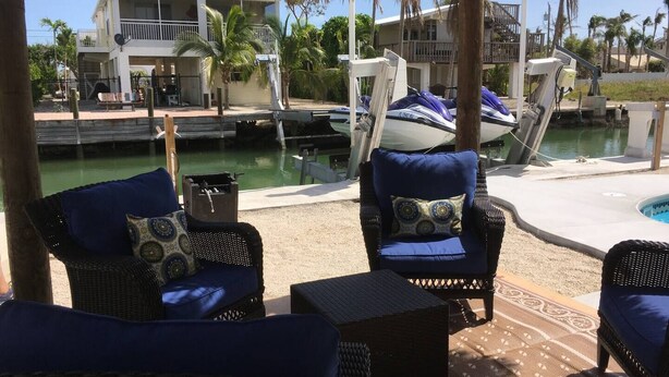 Un espacio de estar techado al aire libre con muebles de mimbre y cojines, con vista a un puerto deportivo con barcos y una piscina.