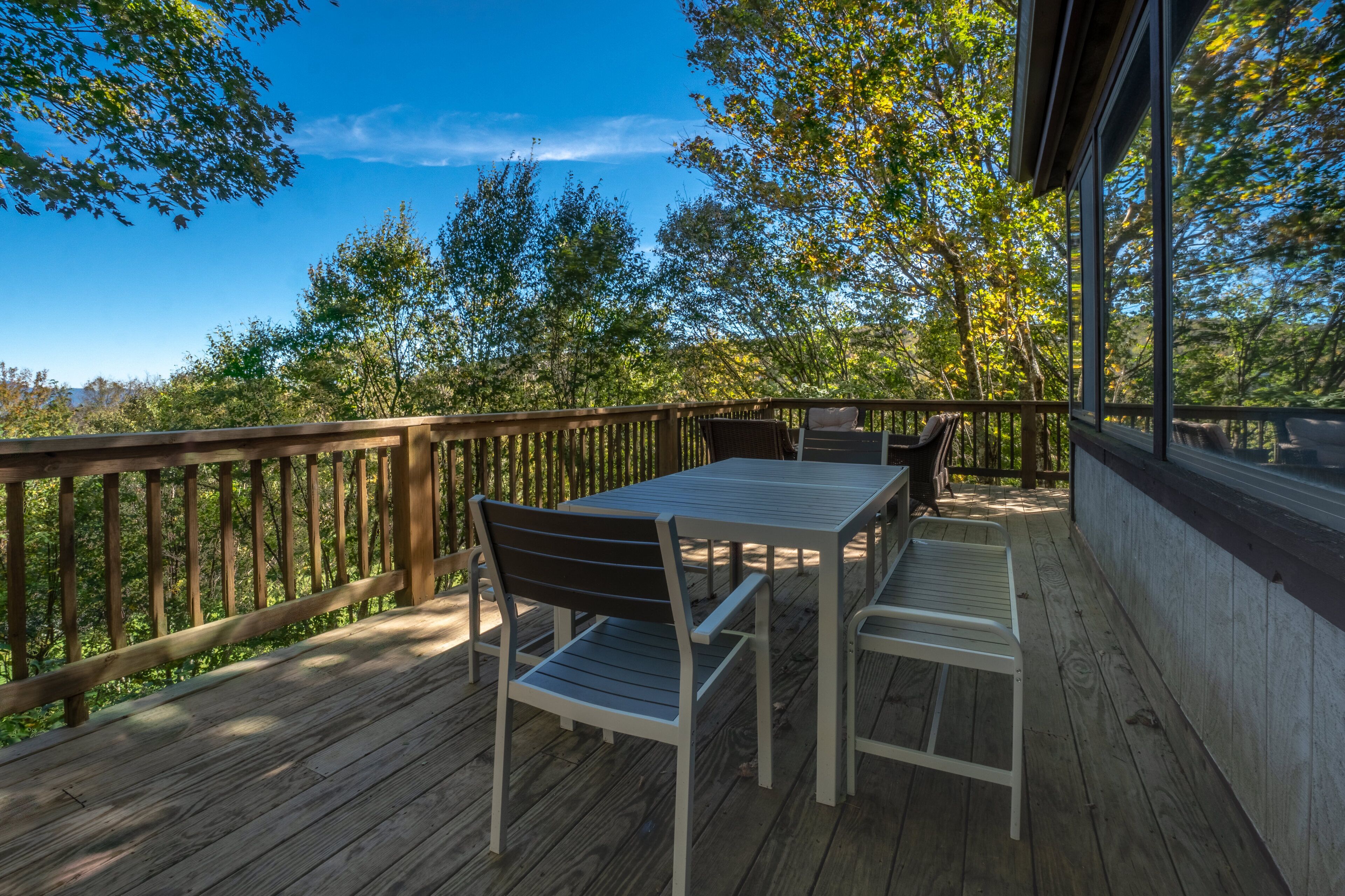 Eine Holzterrasse mit Tisch und Stühlen, umgeben von Bäumen und blauem Himmel.