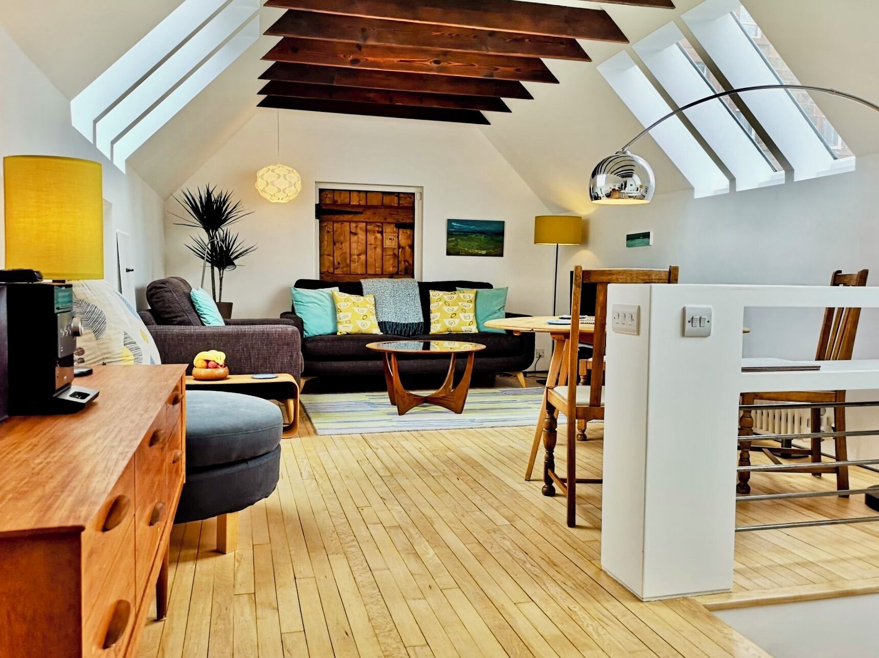 Ein Loft-Wohnraum mit geneigter Decke, Holzbalken, einer Sofagarnitur, einem Esstisch und einer Küchenzeile.