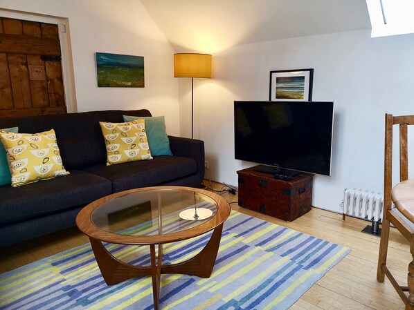 A living room with a dark sofa, yellow patterned pillows, a glass coffee table, a striped rug, a standing lamp, a television on a wooden stand, and a framed picture on the wall.