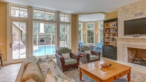 A spacious living room with a large sofa, two armchairs, a coffee table, and a fireplace. There is a pool visible through the glass doors. - 6 Piping Plover-A true smart home with media room! 146 Yard walk to the Beach (Hilton Head Island)