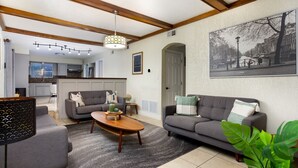 Een moderne woonkamer met een grijze bank, een houten salontafel en een grijs vloerkleed. Achtergrond is een keuken met een raam en een hanglamp.
