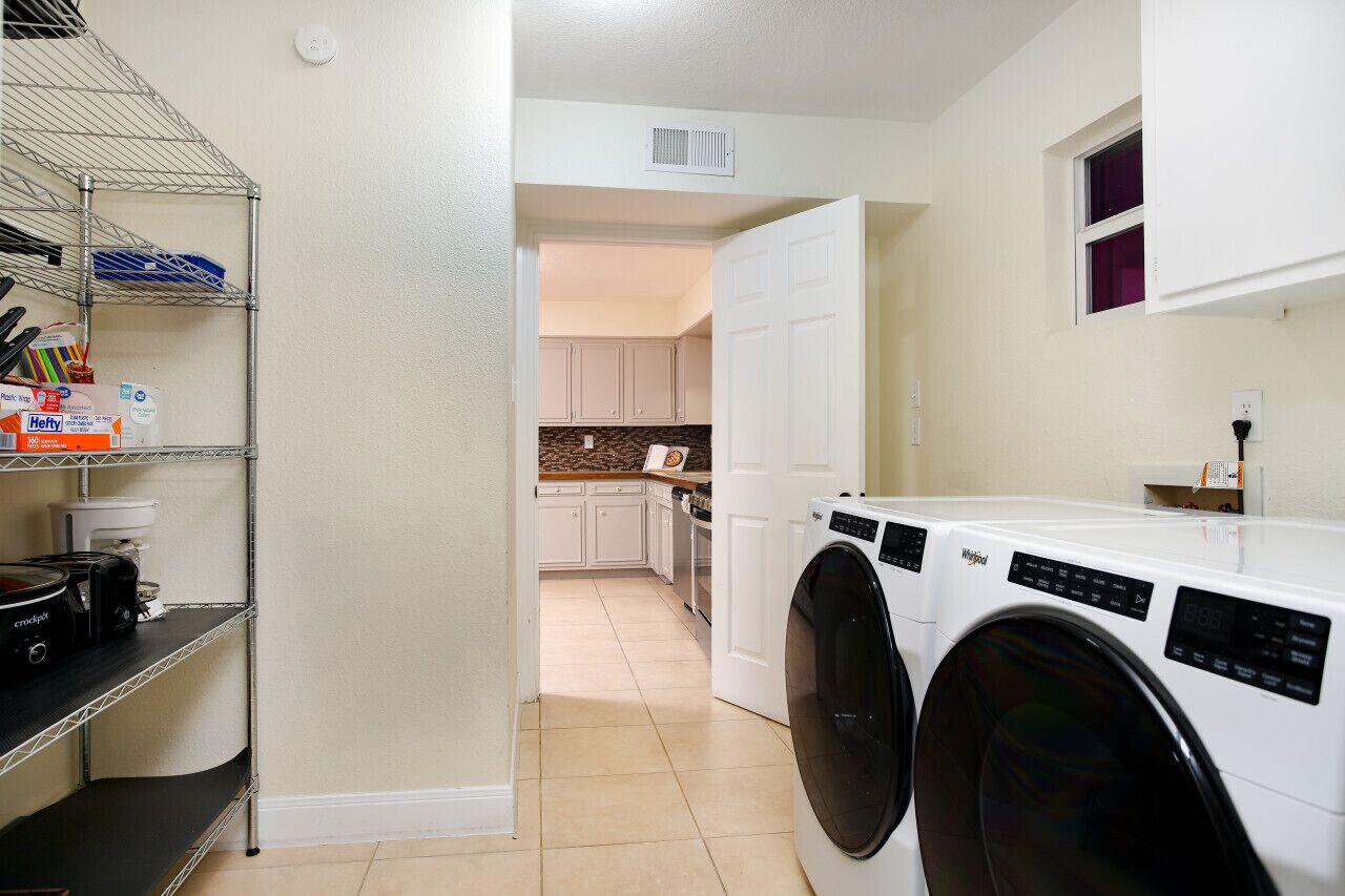 A laundry room with a front-loading washer and dryer, a metal shelf unit with kitchen items, and a kitchen visible in the background.
