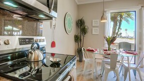 A modern kitchen with a stainless steel kettle on the stove, a digital clock, and a dining area with white chairs and a glass table. - Paradise Retreat | 2BR Bayfront w/ Spacious Patio (San Diego)