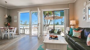 A spacious living room with a dining area, a sofa with decorative pillows, and a view of palm trees and the ocean through large sliding doors. - Paradise Retreat | 2BR Bayfront w/ Spacious Patio (San Diego)