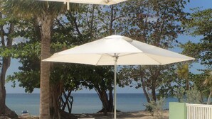 Eine Terrasse direkt am Strand mit einem weißen Sonnenschirm, weißen Stühlen und Blick auf das Meer.