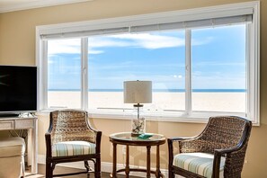 A room with a large window overlooking a beach, two wicker chairs, a small table with a lamp, and a television on a stand.
