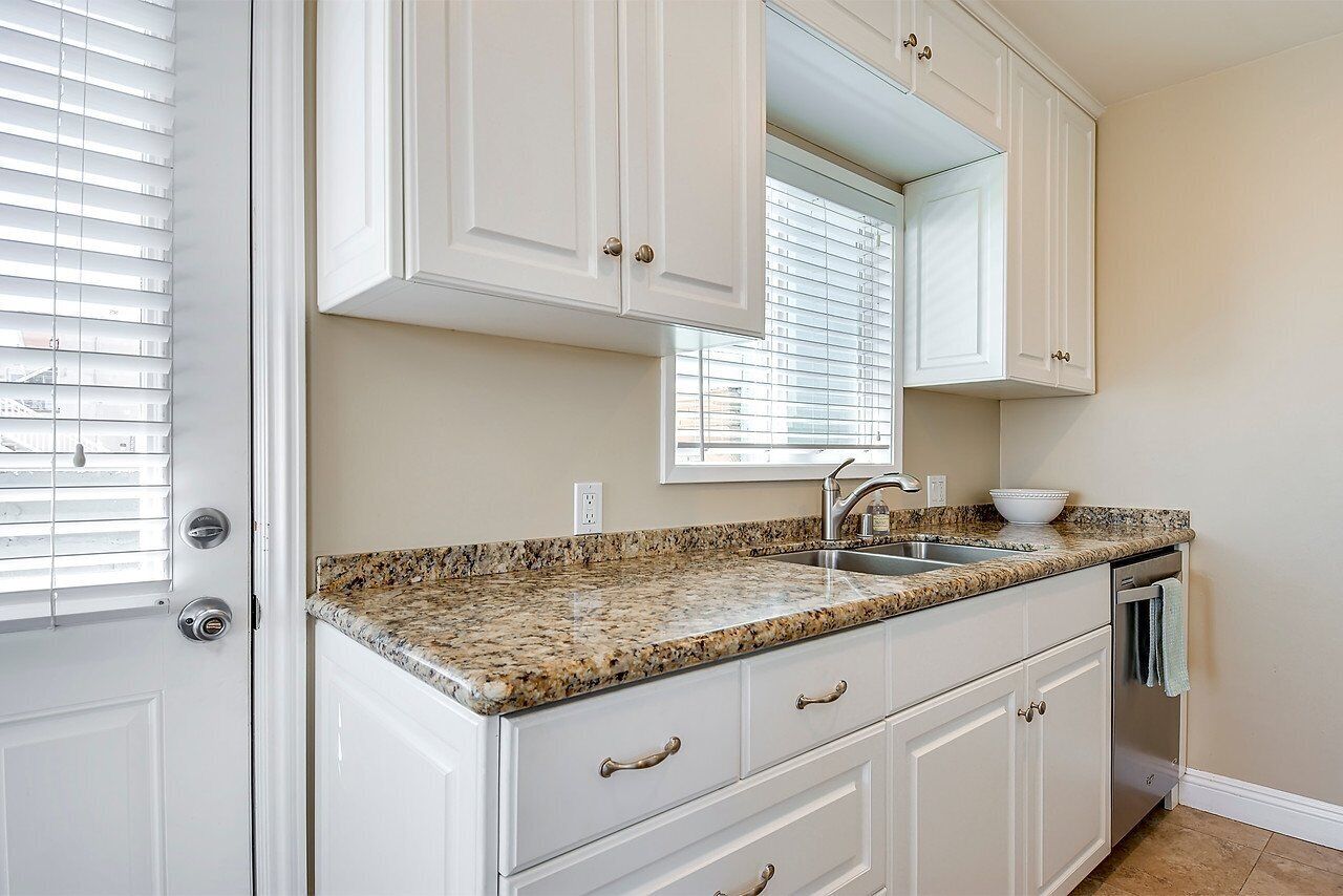 A kitchen with white cabinets, a granite countertop, a sink, and a window with blinds.