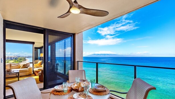 Un balcon avec une table dressée pour le dîner, offrant une vue sur une mer d’un bleu limpide et sur des montagnes lointaines.
