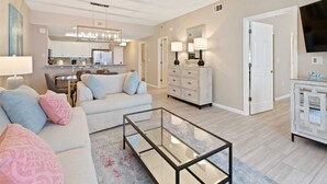 A modern living room with a glass coffee table, a sofa with decorative pillows, a dining area with a kitchen in the background, and a television on the right.