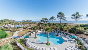 A pool area with lounge chairs and palm trees, surrounded by a fence, overlooking a beach and ocean.